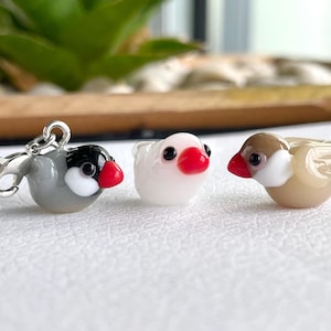 Peut inclure: Trois petites figurines d'oiseaux en verre. Le premier est un oiseau gris avec une tête noire, des joues blanches et un bec rouge, fixé à un fermoir mousqueton en métal argenté. Le deuxième est blanc avec un bec rouge. Le troisième est beige.