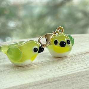 May include: Two small glass bird charms, one with a gold clasp. The birds are green and yellow with white bellies and black eyes.