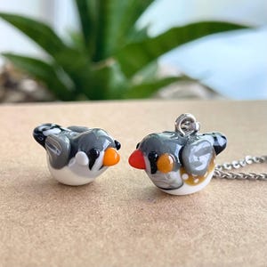 May include: Two small glass bird figurines, one on a silver chain necklace. The birds are grey, white, and orange with black eyes.