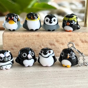 Peut inclure: Une collection de huit petites perles en verre en forme de pingouin fabriquées à la main et un pendentif pingouin sur une chaîne argentée. Chaque pingouin présente un coloris noir, blanc et gris avec des expressions faciales distinctes.