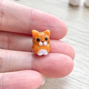 Puede incluir: Una peque&ntilde;a figurita de vidrio de un gato naranja y blanco. El gato tiene ojos negros y una nariz blanca.