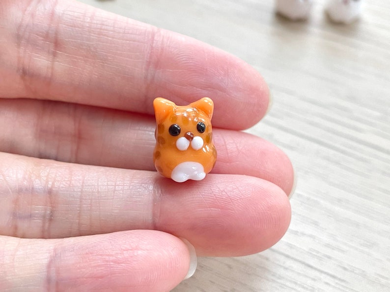 Puede incluir: Una peque&ntilde;a figurita de vidrio de un gato naranja y blanco. El gato tiene ojos negros y una nariz blanca.