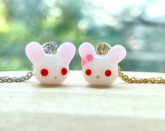 May include: Two white glass bunny charms with red eyes and pink accents on silver and gold chains. The bunny on the left has no bow, while the bunny on the right has a pink bow.