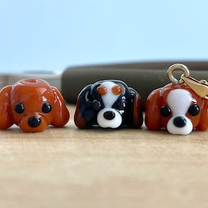 May include: Three glass Cavalier King Charles Spaniel dog charms. One charm is brown and white, one is black and white, and one is brown. The brown and white charm is on a gold chain.