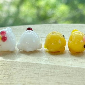 May include: Four glass beads shaped like chicks. Two are white with red and yellow accents, and two are yellow with black eyes and a pink beak.