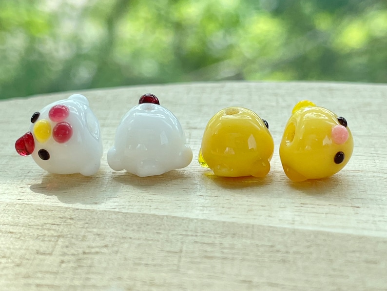 May include: Four glass beads shaped like chicks. Two are white with red and yellow accents, and two are yellow with black eyes and a pink beak.