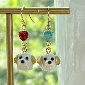 May include: A pair of gold dangle earrings with white dog charms and heart-shaped beads. One earring has a red heart bead and the other has a blue heart bead.