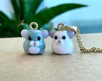 May include: Two glass hamster charms, one gray and one white, with pink accents and black eyes. The white hamster charm is attached to a gold chain.