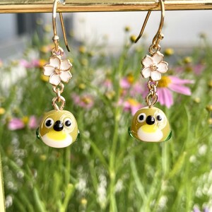 May include: A pair of gold-colored dangle earrings. Each earring features a white flower charm above a small, round, olive-green bird with black eyes and a yellow chest. The earrings are set against a blurred background of flowers.