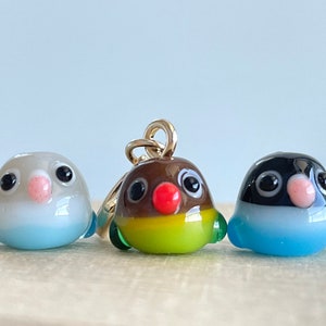 May include: Three glass bird figurines in a row. The birds have black eyes, pink beaks, and colorful bodies. The center bird has a gold loop attached. The birds are white, brown, and black with blue, green, and yellow accents.