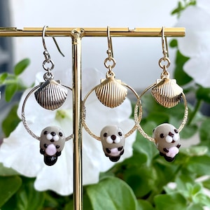 May include: A pair of gold and silver hoop earrings with a seashell charm and a small glass sea otter charm hanging from each hoop.