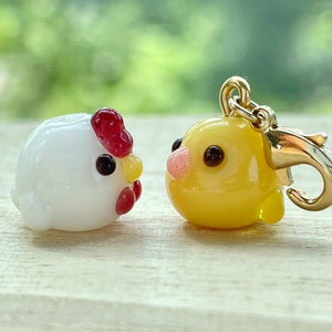 May include: Two small glass animal charms, one white with a red comb and the other yellow. The yellow charm is attached to a gold metal clasp.