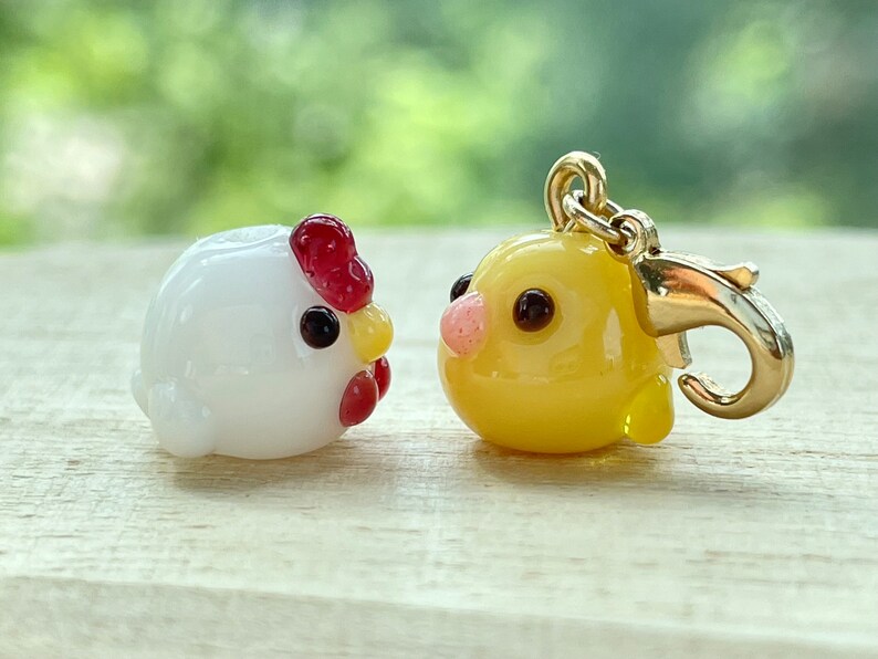 May include: Two small glass animal charms, one white with a red comb and the other yellow. The yellow charm is attached to a gold metal clasp.