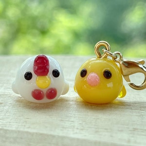 May include: Two small glass figurines of chickens. One is white with red accents and the other is yellow. The yellow chicken has a gold charm attached.