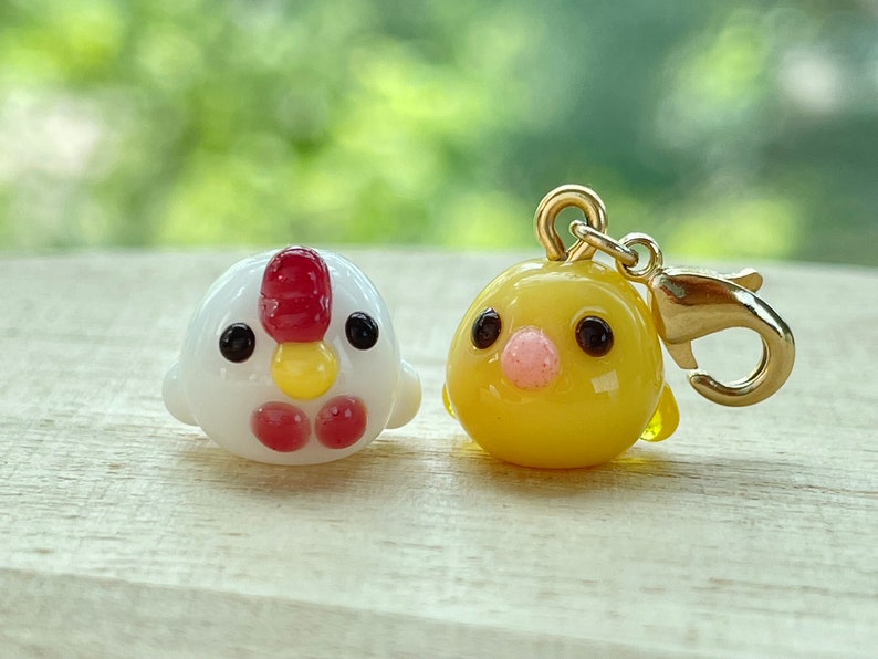 May include: Two small glass figurines of chickens. One is white with red accents and the other is yellow. The yellow chicken has a gold charm attached.