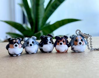 May include: A collection of five small, glass dog figurines in various colors, including black, white, brown, and gray. Each figurine has a round body and a small, detailed face. One figurine is attached to a silver chain, suggesting a pendant.