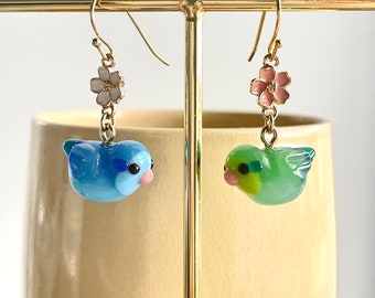 May include: A pair of dangle earrings featuring a blue and green bird charm with a pink or white flower charm above. The earrings are hanging from a gold stand.
