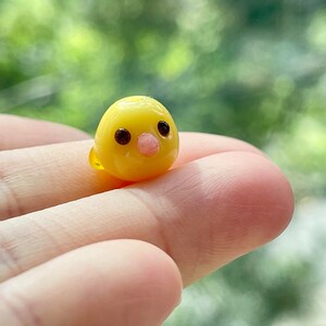 May include: A small, yellow glass figurine of a chick with black eyes and a pink beak.