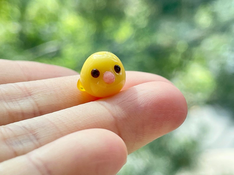May include: A small, yellow glass figurine of a chick with black eyes and a pink beak.
