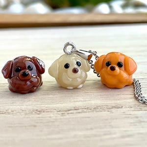 以下が含まれることがあります： 滑らかで光沢のある素材で作られた、犬の形をした3つの小さなチャーム。1つはダークブラウン、1つはクリーム色、もう1つはオレンジ色です。クリーム色のチャームにはシルバーカラーの金属製バチカンが付いており、細いシルバーカラーの金属チェーンに繋がっています。