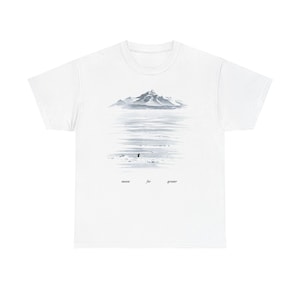 May include: White t-shirt featuring a serene landscape design. The graphic depicts a mountain range reflected in water, with a small figure in the foreground. The text "meant for greater" is printed below the image.