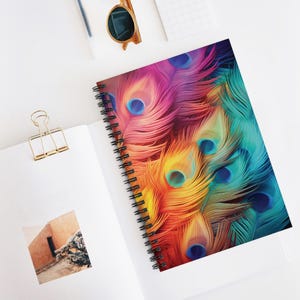 May include: A spiral-bound notebook with a vibrant peacock feather design. The cover features a close-up of colorful feathers in shades of pink, orange, yellow, and teal. The notebook is open, revealing blank white pages and a gold paper clip.