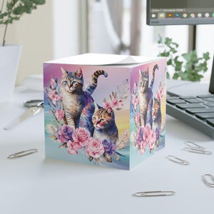May include: A square notepad with watercolour illustrations of cats and flowers. The cats have blue and brown fur. The background has a gradient of pink, purple, and blue colours.