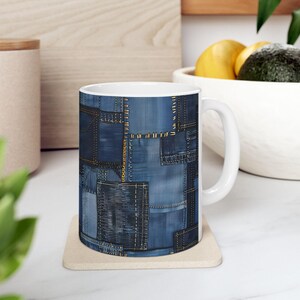 Puede incluir: Taza de cerámica blanca con un diseño de patchwork de mezclilla. La taza muestra varios tonos de parches de mezclilla azul cosidos con hilo dorado, creando un patrón texturizado. La taza tiene un asa estándar.