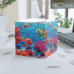 May include: A cube-shaped notepad featuring a vibrant underwater scene. The design showcases colorful fish swimming amidst coral reefs in shades of red, orange, and blue. The top of the notepad is white, providing a contrast to the aquatic illustration.