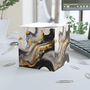 May include: A square notepad holder with a white top and a swirling abstract design in black, gold, and white. The holder sits on a white desk with scattered silver paper clips, near a computer and a plant.