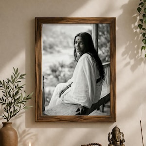 Puede incluir: Fotografía en blanco y negro enmarcada de una mujer con un sari blanco, sentada en un porche. El marco de madera rodea la imagen. Una estantería de madera debajo contiene un jarrón con vegetación, una vela encendida, un libro, una estatua e incienso.