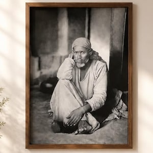 May include: A framed monochrome portrait of a person seated on the floor, wearing a head covering and loose clothing. The portrait is in a dark wooden frame. The image is displayed on a light-coloured wall.