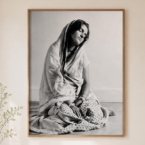May include: Monochrome framed print featuring a person seated on the floor, wrapped in cloth. The person wears a head covering and appears contemplative. The print is displayed in a light wood frame, suitable for home decor.