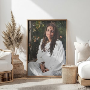 Puede incluir: Una mujer vestida con una túnica blanca se sienta frente a un fondo verde frondoso. Tiene el pelo largo y oscuro y lleva un pequeño punto rojo en la frente.