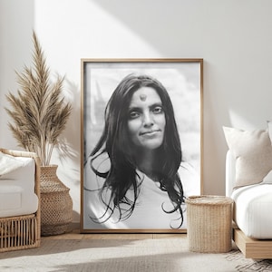 Puede incluir: Retrato enmarcado en blanco y negro de una mujer con cabello largo y oscuro y un bindi. El retrato se exhibe en una habitación con tonos neutros, incluyendo un jarrón tejido con plantas secas y una mesa auxiliar de mimbre.