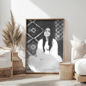 Puede incluir: Fotografía en blanco y negro de una mujer sentada en una postura meditativa. Lleva una túnica blanca y tiene los ojos cerrados. La foto está enmarcada en un marco de madera.