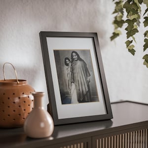 Puede incluir: Una fotografía en blanco y negro enmarcada en negro de dos personas de pie juntas. Las personas visten ropa tradicional. La fotografía está sobre una superficie de madera marrón oscuro.