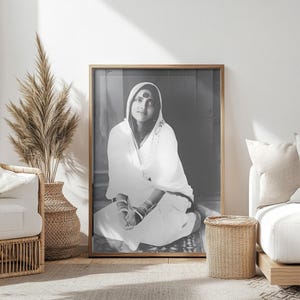 Puede incluir: Una fotografía en blanco y negro de una mujer vestida con una túnica blanca y sentada con las piernas cruzadas. Lleva un bindi en la frente y tiene una expresión serena en su rostro.