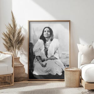 Puede incluir: Fotografía en blanco y negro de una mujer con una túnica blanca sentada con las piernas cruzadas. Lleva un bindi en la frente y un collar alrededor del cuello.