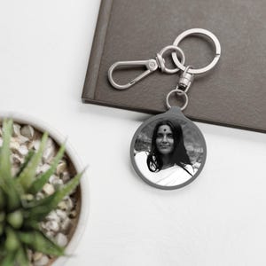 May include: A silver keychain with a round, gray photo charm featuring a black and white image of a woman with long hair. The woman is wearing a white garment and has a small red dot on her forehead.