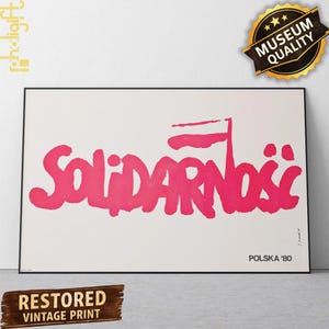 Puede incluir: Una impresión rectangular enmarcada que presenta la palabra SOLIDARNOŚĆ en letras gruesas de color rosa brillante con estilo de pincelada sobre un fondo blanco liso. El texto POLSKA '80 aparece en letras negras pequeñas en la esquina inferior.