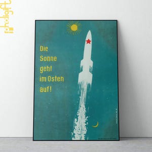 以下が含まれることがあります： 水色の背景に、白いロケットと赤い星、黄色い太陽と三日月が描かれた、額入りのヴィンテージスタイルのポスター。ドイツ語のテキスト「Die Sonne geht im Osten auf！」は黄色で表示されています。