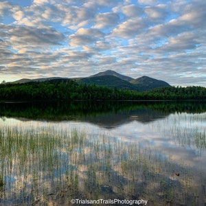 Puede incluir: Un lago sereno que refleja un cielo nublado y una cordillera en la distancia. El agua está tranquila y el reflejo de las nubes es claro.