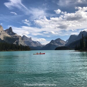 May include: Two people in a red kayak paddle across a turquoise lake with mountains in the background. The sky is blue with white clouds.