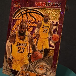 May include: A rectangular NBA Hoops trading card featuring LeBron James in a yellow Los Angeles Lakers jersey with the number 23. The card has a purple glitter border, a gold textured background, and includes the text NBA HOOPS and LEBRON JAMES.