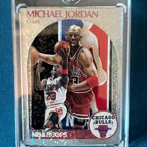 May include: A Michael Jordan NBA Hoops trading card encased in a clear plastic magnetic holder. The card features a die-cut design with two images of the player in a Chicago Bulls uniform, the text Michael Jordan, Guard, and a Chicago Bulls logo.