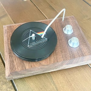 Könnte beinhalten: Ein Holz-Untersetzer-Set, das einem Miniatur-Plattenspieler nachempfunden ist. Das Set enthält einen Stapel schwarzer Schallplatten-Untersetzer mit dem Albumcover "Dark Side of the Moon" von Pink Floyd, einen silberfarbenen Tonarm und zwei silberne Knöpfe.