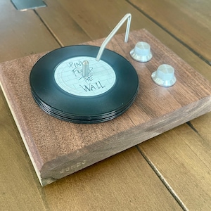 May include: A miniature record player coaster with a stack of black vinyl records. The record label reads "Pink Floyd The Wall". The coaster sits on a wooden base with two silver knobs and a silver tone arm.