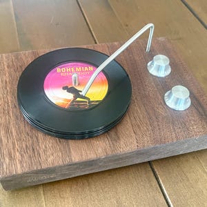 May include: A wooden coaster set designed to resemble a record player. The set includes a stack of black vinyl coasters with the words "Bohemian Rhapsody" on the label, a metal tone arm, and two silver knobs.