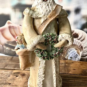 May include: A detailed figurine of Santa Claus, wearing a cream-colored robe with white trim and a matching hat. He holds a green wreath with red berries and a small doll in a sack. The figurine is on a wooden surface.